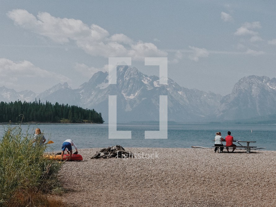 people sitting at a picnic table in front of a mountain lake 