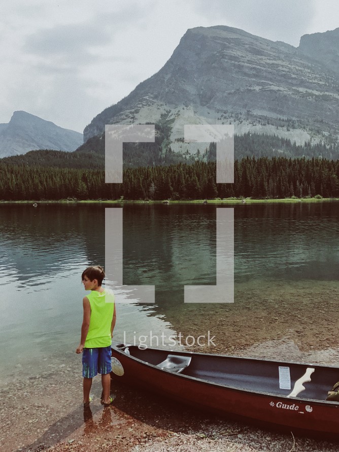 child standing by a boat on a lake shore 