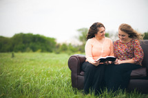 Smiling mother and daughter reading the Bible together while sitting on a sofa in a field.