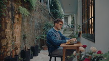 Young male freelancer drinking tea, typing on laptop, working remotely in a small outdoor area beside house in the evening. Medium full shot
