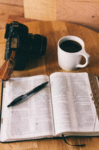 An open Bible, cup of coffee, and a camera on a table.