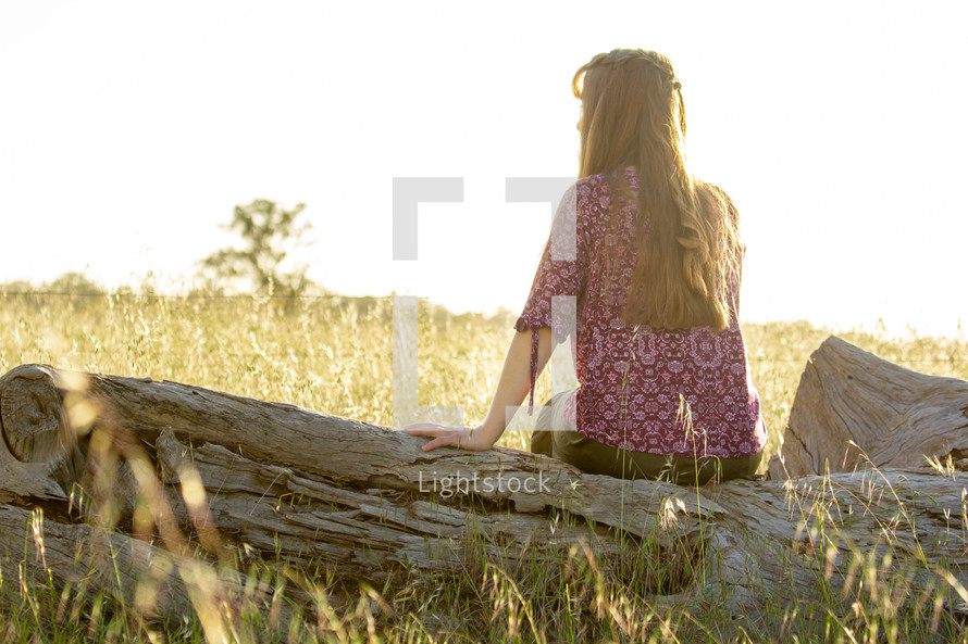 a teen girl sitting on a log in a field alone 