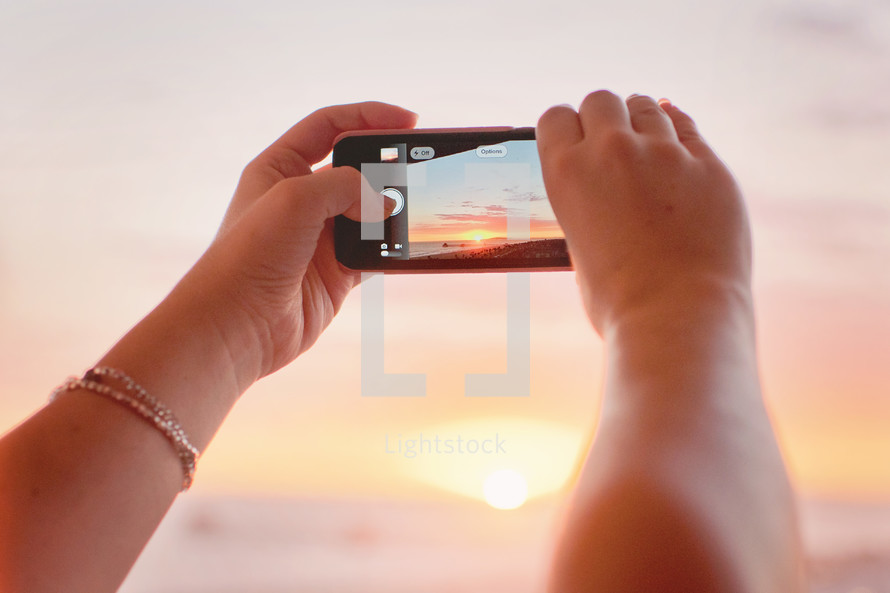 a woman taking a picture of the beach at sunset with her cellphone 