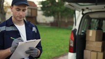Young delivery man checking delivery list. Delivery man in cap and documentation in hands outdoor. Courier with parcel. Male handsome courier checks the parcels against the delivery list. Trunk with parsels.