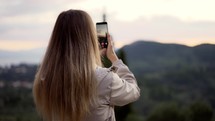 Blonde woman takes a photo of mountains with a smartphone in her hands.