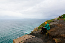 a woman in a raincoat sitting on a shore on the Cliffs of Moher 