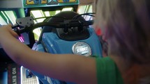 Six-year-old girl riding a motorcycle in an arcade, pressing the start button and navigating through the game, viewed from behind and above