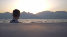 Little boy looking out at an ocean with mountains in the background during sunset.