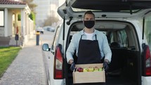 Friendly smiling man in mask holding wooden box with apples at street outdoors, portrait of happy deliveryman in apron and gloves standing near the trunk. Online shopping delivering, smart vegetables express delivery service, slow-motion.