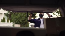 Delivery service. Delivery man in a protective mask and gloves opening the trunk and taking cardboard box parcel. Man closing minivan trunk with parcels. Footage from the backseat of the car.