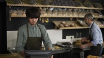 Young cashier in apron typing on touch screen, handsome bakery worker slicing bread in the background.