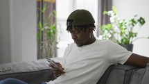 Black African American man using smartphone while resting on a sofa in living room.