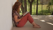 Woman sitting outside, leaning against a wall, reading a book.