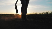 Farm agriculture concept. Farmer with a shovel in rubber boots walks across field at sunset. Silhouette man gardener walking along the road along the field. Tired man after hard work on the ground.