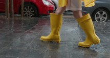 Child's feet in tall yellow rubber boots walking on a wet sidewalk, passing by moving cars during heavy rain