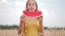 Cute girl eating juicy watermelon standing in wheat field on sunny summer day. Child eats a delicious watermelon, summer lifestyle. Kid emotion at summer vacation.