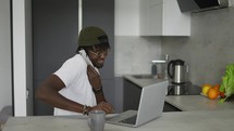 Black man working at home in kitchen talking on smartphone and using laptop.