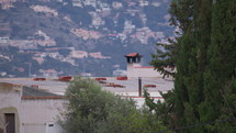 Behind the evergreen trees, smoke is coming out of the chimney on the roof of a white house situated below the mountain. On the mountain, there are white houses of the city in Spain