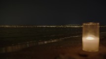A flickering candle in a glass jar, blurry in the foreground on a beach at night, with the clear, distinct lights of a coastal city in the focused background.