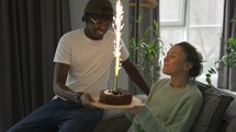 A husband brought the birthday cake with big sparkling candle to his wife in the living room.
