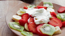 A close-up of a person's hand spooning Greek yogurt onto a fruit salad made with strawberries, kiwis, and bananas