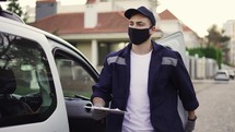 Courier going out from the van and takes cardboard box package out of delivery car makes notes in delivery list. Courier in mask, cap and gloves on the way to deliver postal parcel to a client.