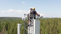 Skyward Ascent: Drone View of Telecom Technicians Scaling Forest Tower Heights