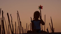 A lone girl on the beach thoughtfully constructs a bamboo enclosure, placing a colorful wind spinner on a bamboo straw under the softly fading sunset glow