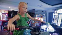 A blue-eyed blonde girl, 6 years old, at an arcade entertainment center. She sits at the helm of a motorcycle. The motorcycle jerks from side to side