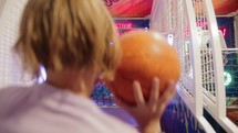 Teenage boy with long hair, viewed from the back, throws basketballs into a moving hoop at an indoor amusement arcade