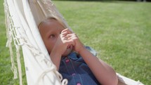 A child lies in a hammock, her gaze thoughtful and distant, amidst the bright greens of an outdoor setting