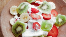 A plate with a salad made from fresh fruits, including strawberries, bananas, and kiwis, beautifully arranged. It is dressed with greek yogurt and sprinkled with seeds on top