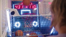Teenager with long hair, shot from behind, throwing basketballs into a moving hoop at an entertainment game center