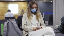 Woman tourist wearing medical protection mask using mobile phone in airport terminal.
