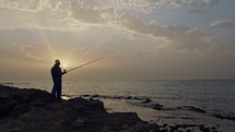 Old fisherman standing on sea side rocks and fishing against the sunset