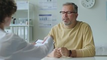 Mid-Aged Man Giving Medical Record and Talking to Doctor in Clinic
