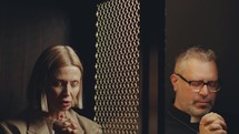 Senior female parishioner and priest sitting on both sides of lattice screen and praying to absolve sins during confession in Catholic church
