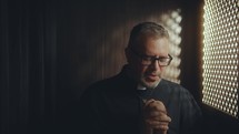 Clergyman with glasses and grey hair clasping hands in prayer inside dimly lit confessional booth illuminated by perforated screen light
