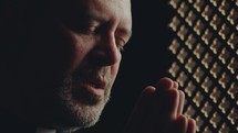 Priest sitting in confessional booth and saying prayer with hands clasped and eyes closed. Close-up view
