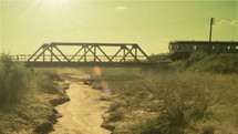 The silhouette of a commuter train traveling over a an older bridge on a sunny day.