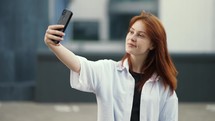 Red-haired girl in a shirt makes a selfie on a city background.