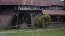 A wide shot of a historic wooden building with a large watermill wheel, surrounded by greenery, with a stone base and wooden details