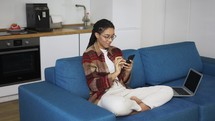 Young woman using smartphone at home on a couch with laptop.