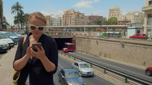 A young woman in white-framed sunglasses stands on a busy street next to a tunnel, holding a mobile phone in her hands