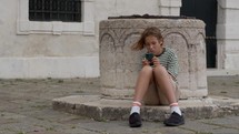 A teenager in a striped shirt sits on a Venice square, engrossed in his phone, leaning against an architectural feature
