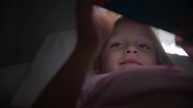 Blonde girl, six years old, lying in bed, plays on the phone, smiles at the game. Reflections on her face from the phone