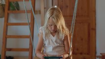 A concentrated young girl browses her smartphone while seated on a rope swing in a home setting