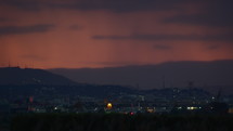 Against the backdrop of a red sunset sky, small houses of a suburban town and passing cars with rain in the far background