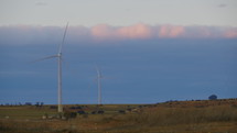 Two wind turbines with electric generators - located on the left side of the frame - at sunset in a field