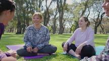 Group of positive mature women sitting on fitness mats on green lawn in the park, smiling and discussing something when having a rest after outdoor yoga practice